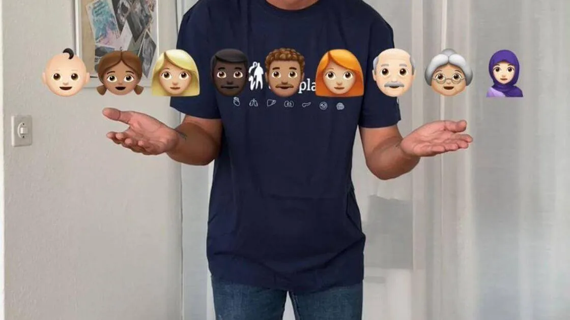 Man in a navy T-shirt printed with organ icons holds out open palms beneath a row of diverse face emojis