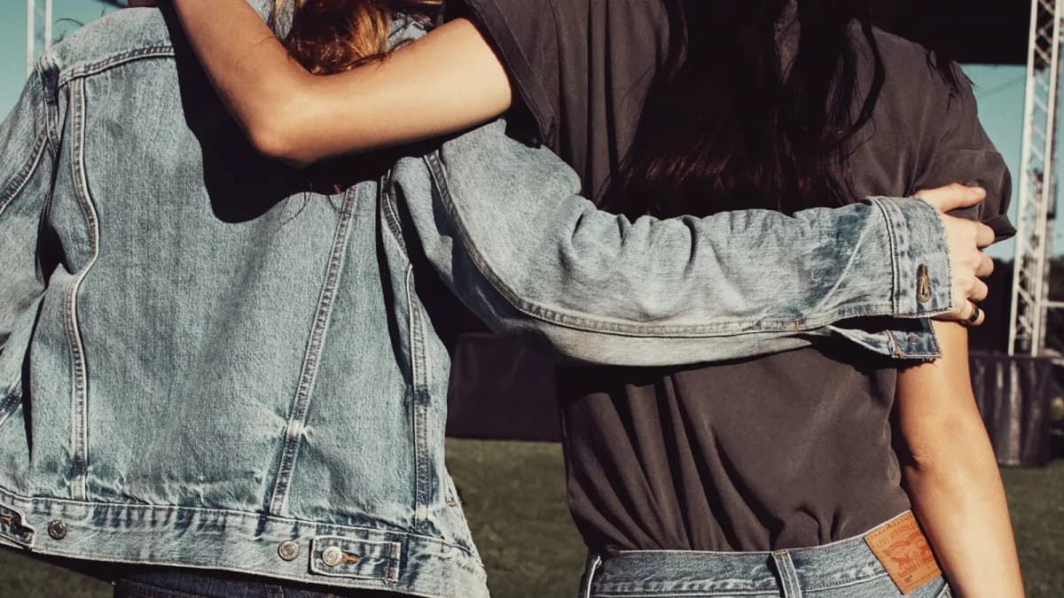 Two people seen from behind with arms around each other, one in a light denim jacket, by an outdoor stage on grass