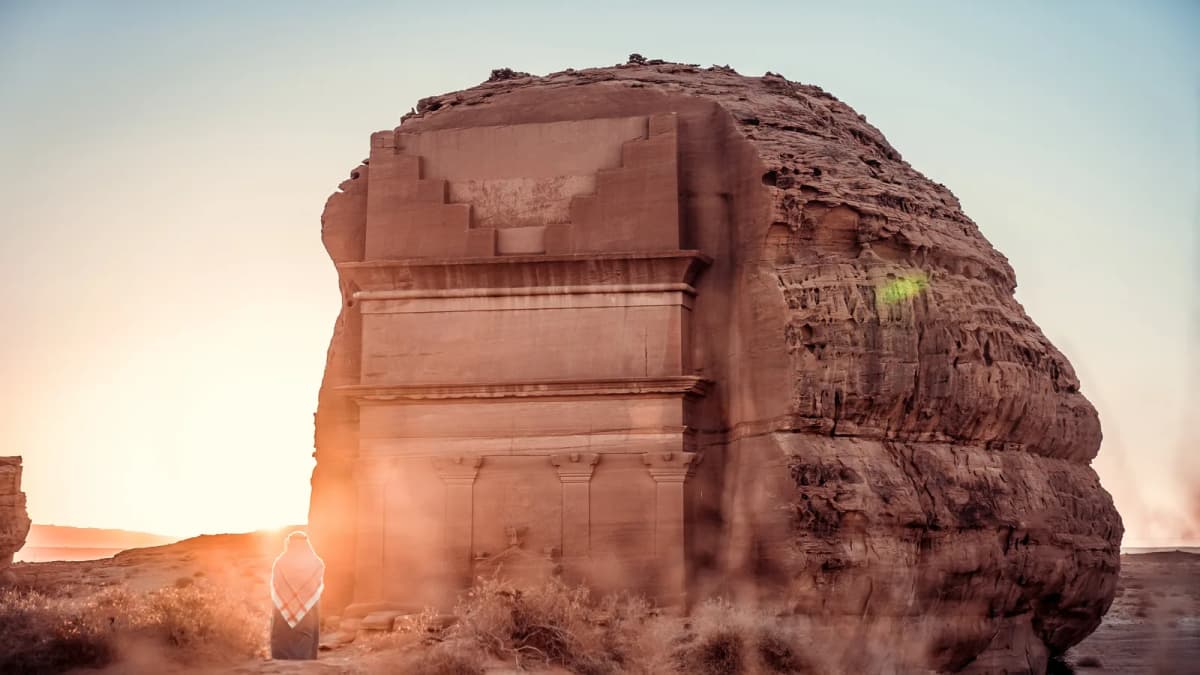 Person in a red-checked headscarf stands before a huge sandstone rock with a tomb façade carved into it, desert at sunset