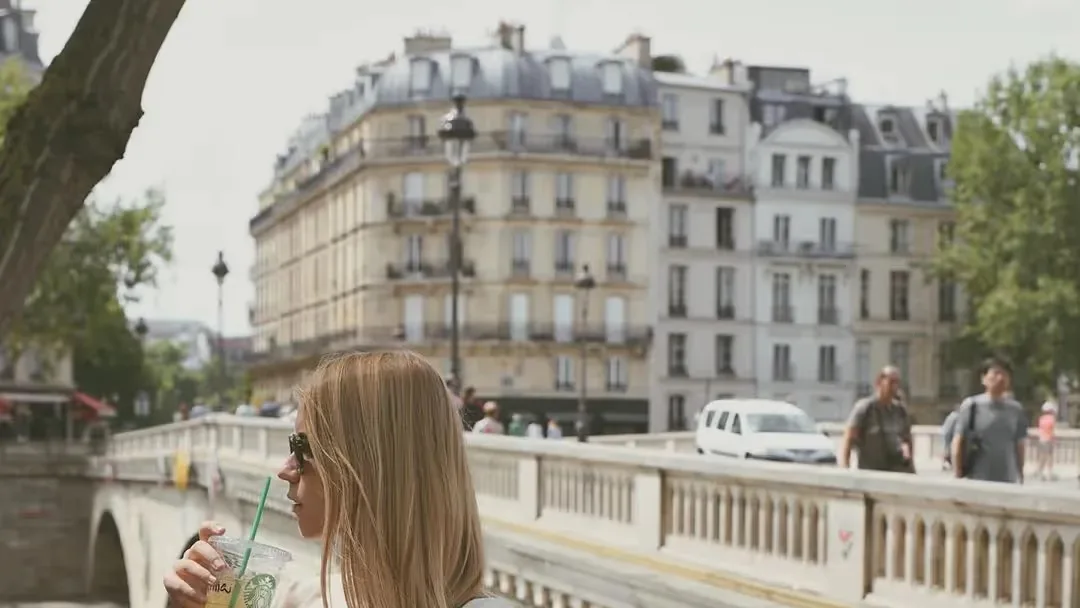 Woman with long blonde hair and sunglasses sips an iced Starbucks drink by a stone bridge, with cream city buildings behind