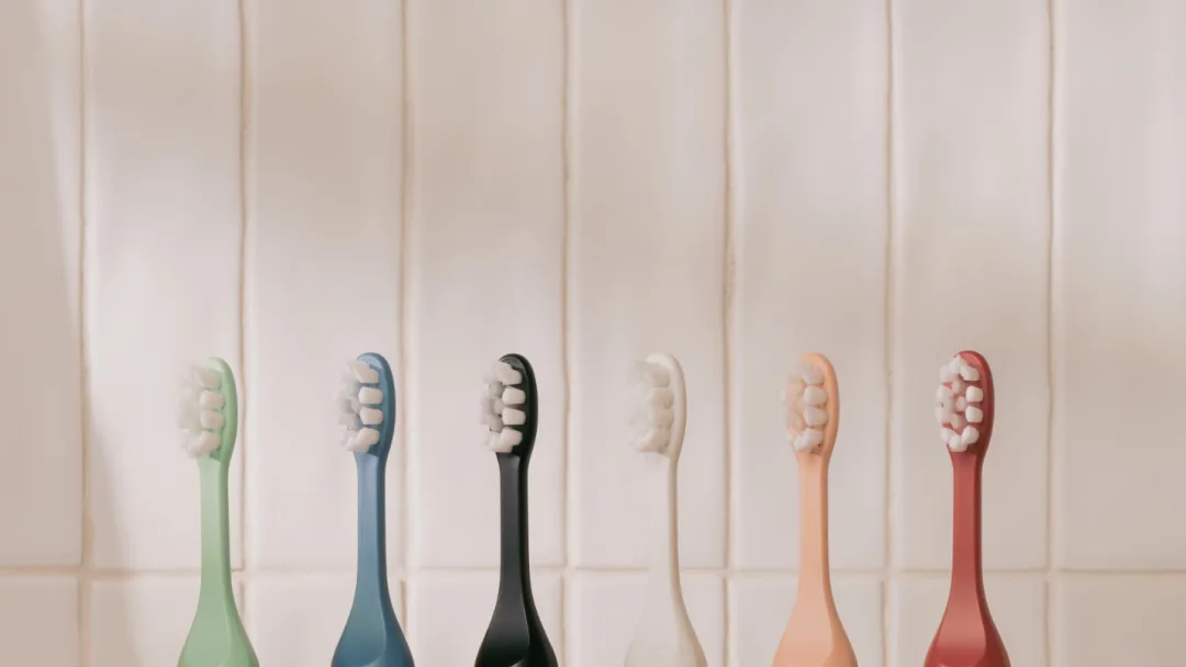 Six SURI electric toothbrush heads in green, blue, black, white, peach and red lined up against a white tiled wall