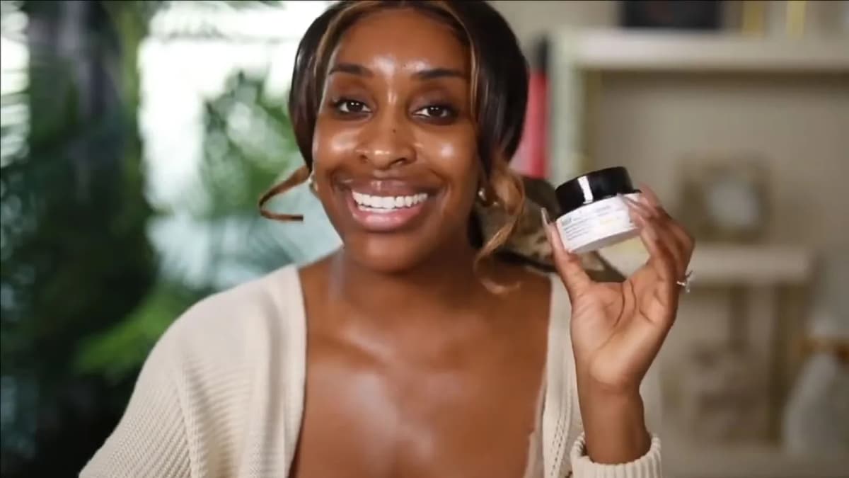 Smiling woman in a cream cardigan holds up a small white belif cream jar with a black lid in a softly lit room with plants