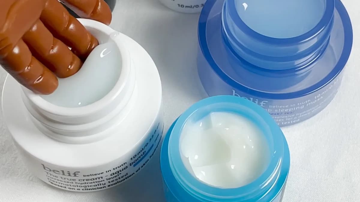A doll hand dips into an open belif "The true cream - aqua bomb" jar beside two open blue jars on white cloth