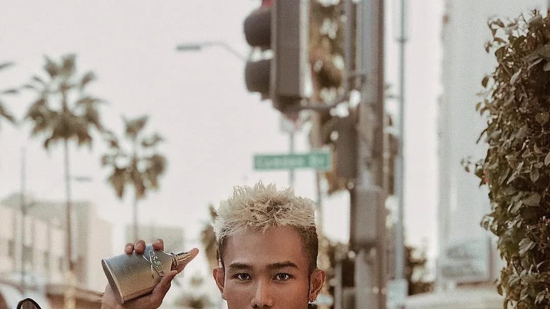 Man with bleached spiky hair holds up a silver perfume bottle on a palm-lined city street beside a traffic light
