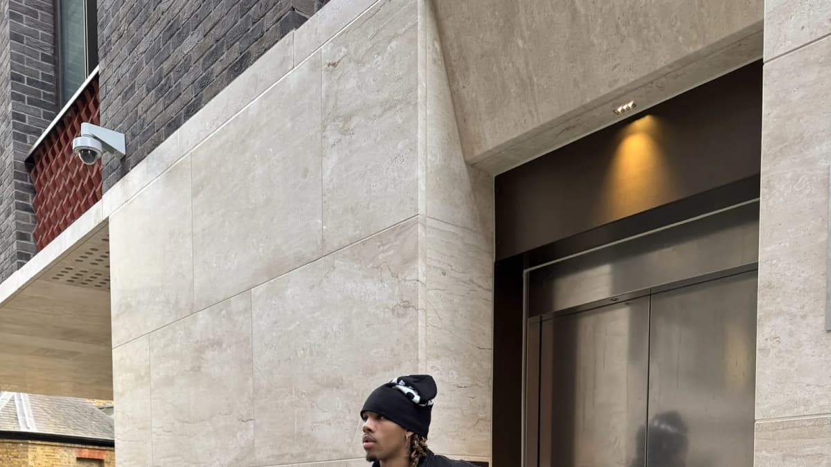 MUSERO, Tay Moses in a black beanie, jacket and wide trousers outside a lift entrance
