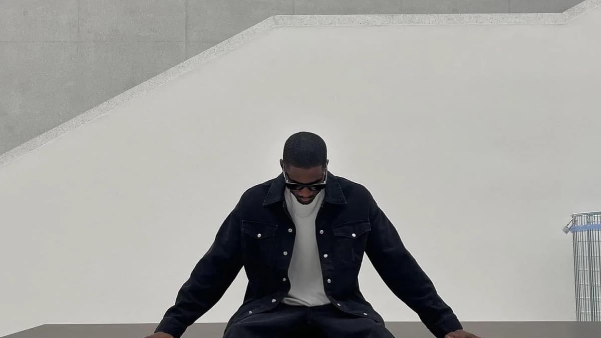 Calvin Klein, Pierre Laurent in a black denim jacket and sunglasses sitting on a bench, legs stretched out