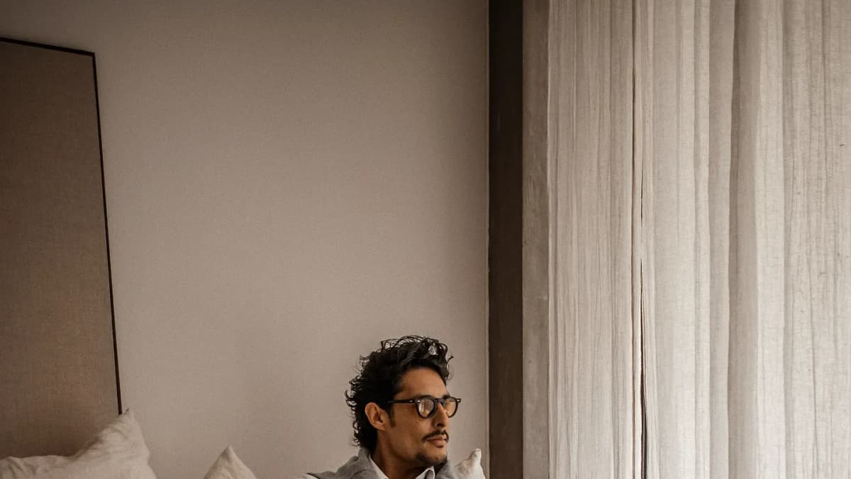 Man with curly dark hair and glasses in a gray sweater sits among white pillows, looking toward linen curtains