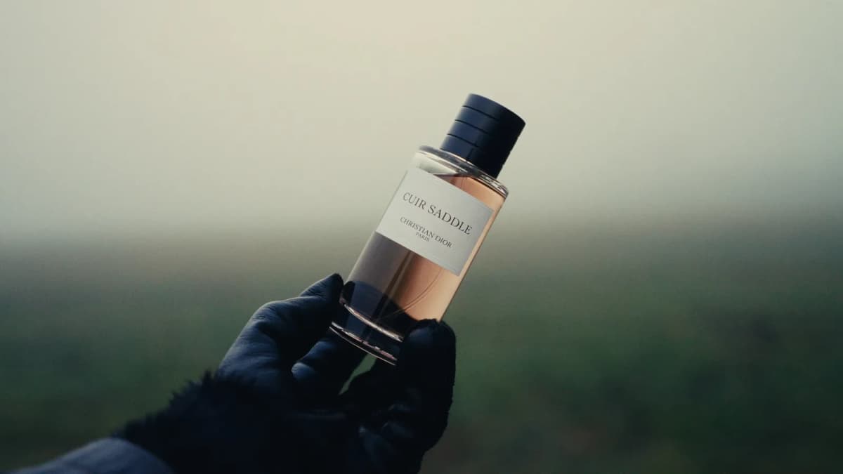 Black-gloved hand holds a Dior Beauty perfume bottle labeled "CUIR SADDLE CHRISTIAN DIOR PARIS" against a misty field