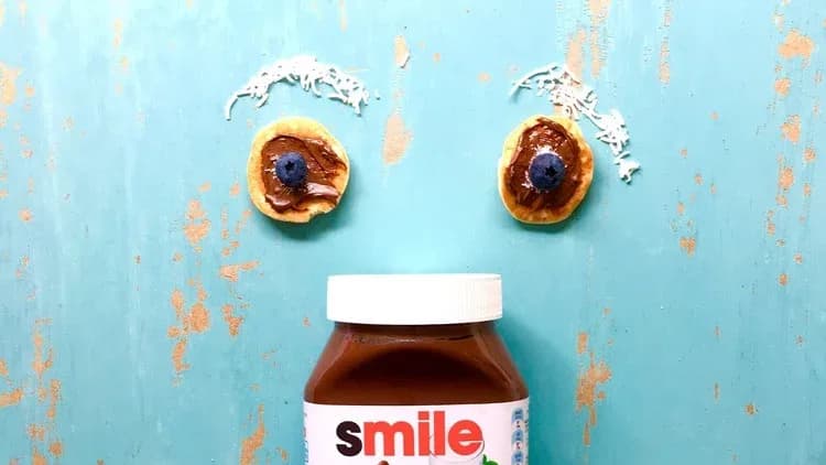 Nutella jar labeled "smile" below two spread-topped mini pancakes with blueberry eyes and coconut brows, on teal wood