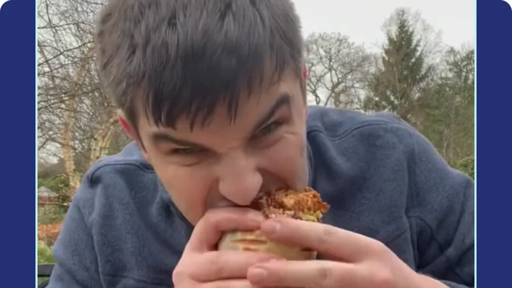 Young man with dark hair in a blue fleece takes a big bite of a grilled wrap outdoors, with bare trees behind him
