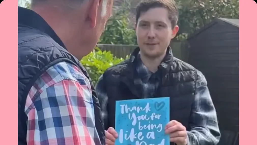 Young man in a garden holds up a blue card reading "Thank You for being like a Dad" to an older man in a plaid shirt