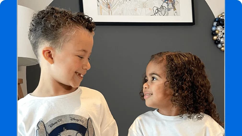 Smiling boy in a "Disney 100" T-shirt and a curly-haired girl in white look at each other in front of a gray wall