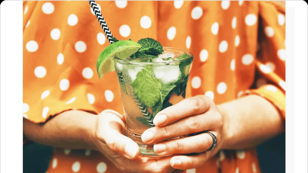 Person in an orange polka-dot top holding a mojito with mint, ice, a lime wedge and a striped straw in both hands