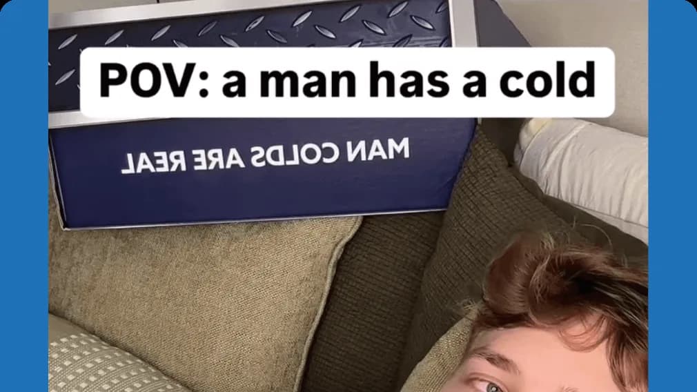 Young man lying on a couch by a navy box reading "MAN COLDS ARE REAL", with caption "POV: a man has a cold"