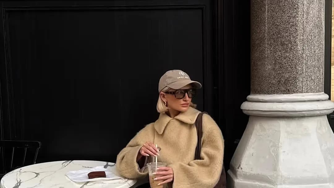 Blonde woman in a beige cap, sunglasses and fuzzy camel coat sits at a marble café table holding an iced coffee