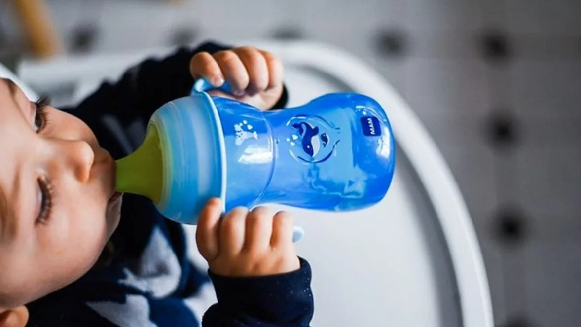 Toddler in a high chair drinks from a blue MAM sippy cup with a whale design and green spout, holding it with both hands