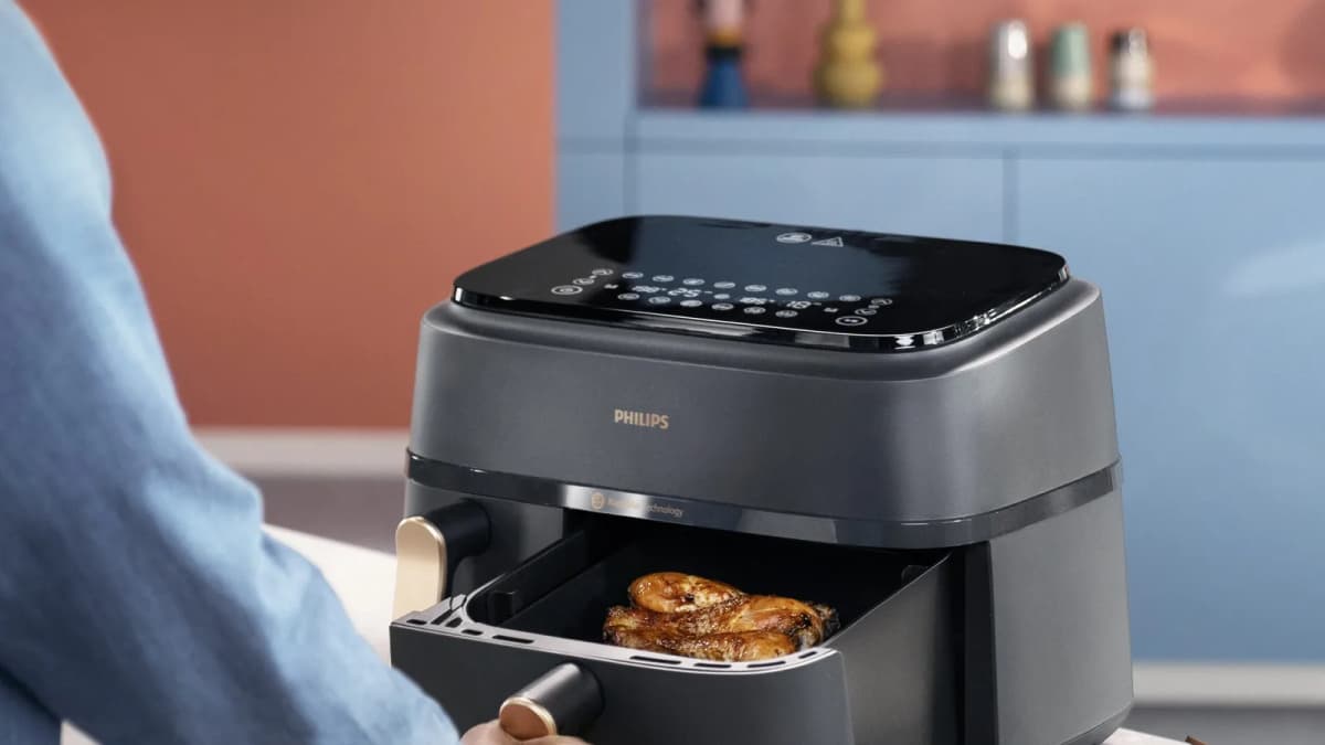 Person in a blue shirt pulls out the basket of a dark grey Philips airfryer, showing roasted chicken, in a colorful kitchen