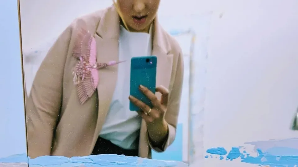 Woman in a beige blazer with a pink beaded bird brooch takes a mirror selfie with a teal smartphone