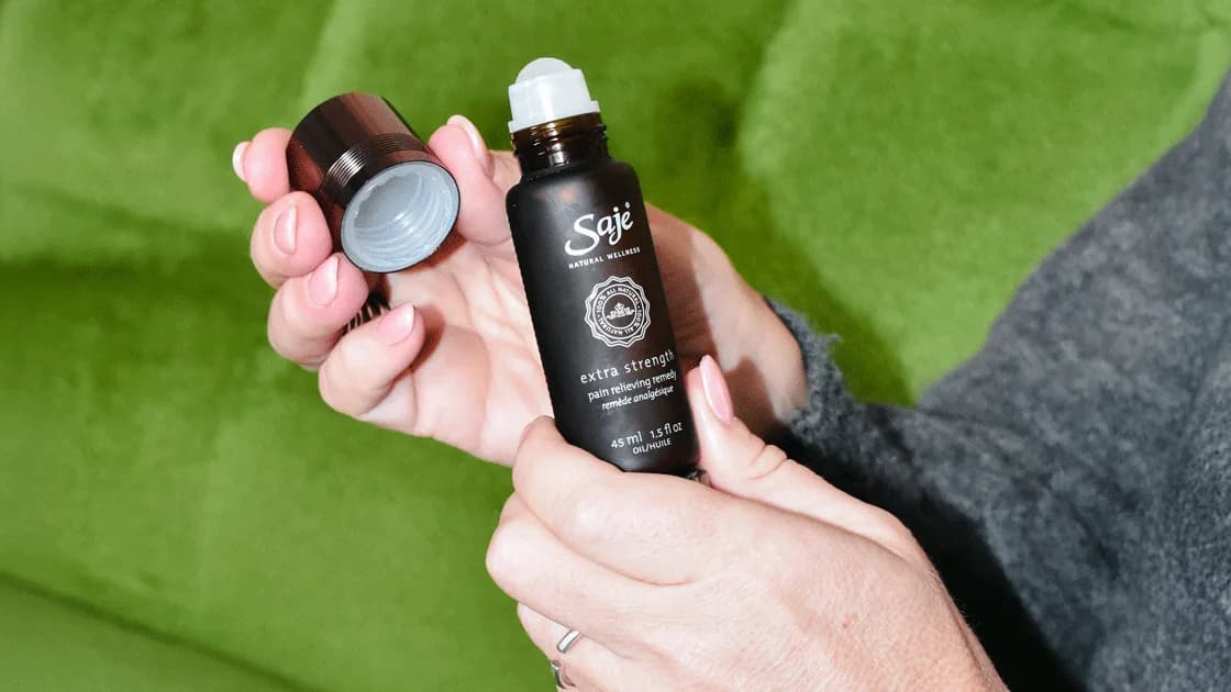Hands hold an open Saje roll-on bottle labeled "extra strength pain relieving remedy" and its cap over a green sofa