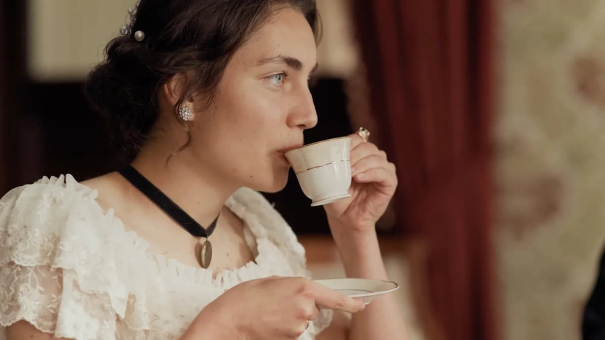 Woman in a white lace period dress and black velvet choker sips from a china teacup holding a saucer in a vintage room
