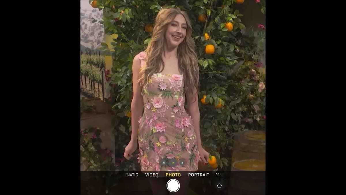 Phone camera view of a smiling woman with long wavy hair in a floral mini dress posing before an orange tree, "PHOTO" mode