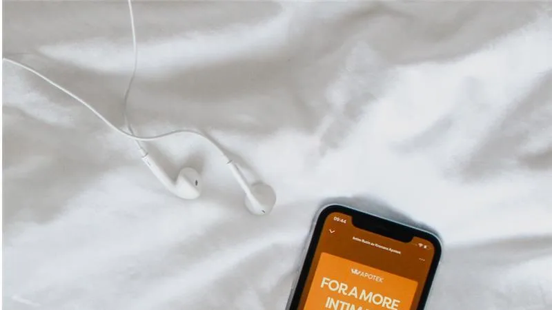 Wired white earphones and a phone on white bedsheets, screen showing a Kronans Apotek audio cover reading "FOR A MORE"