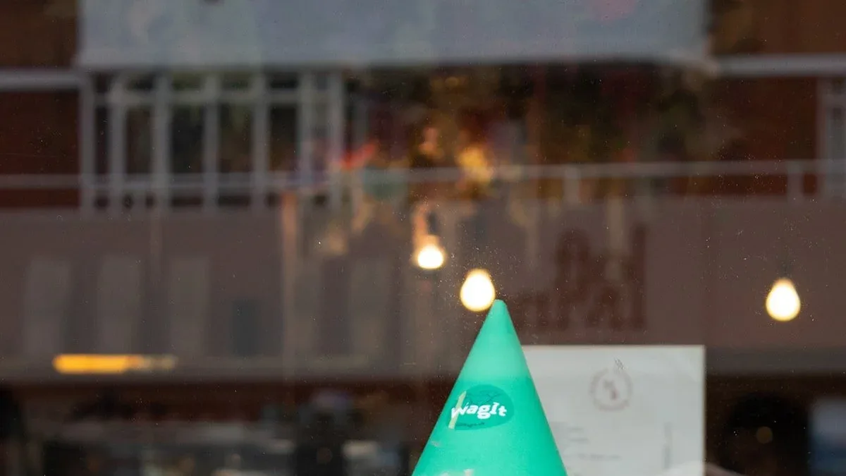 Green party hat with a round "wagit" logo seen through a shop window reflecting a brick building and glowing bulbs