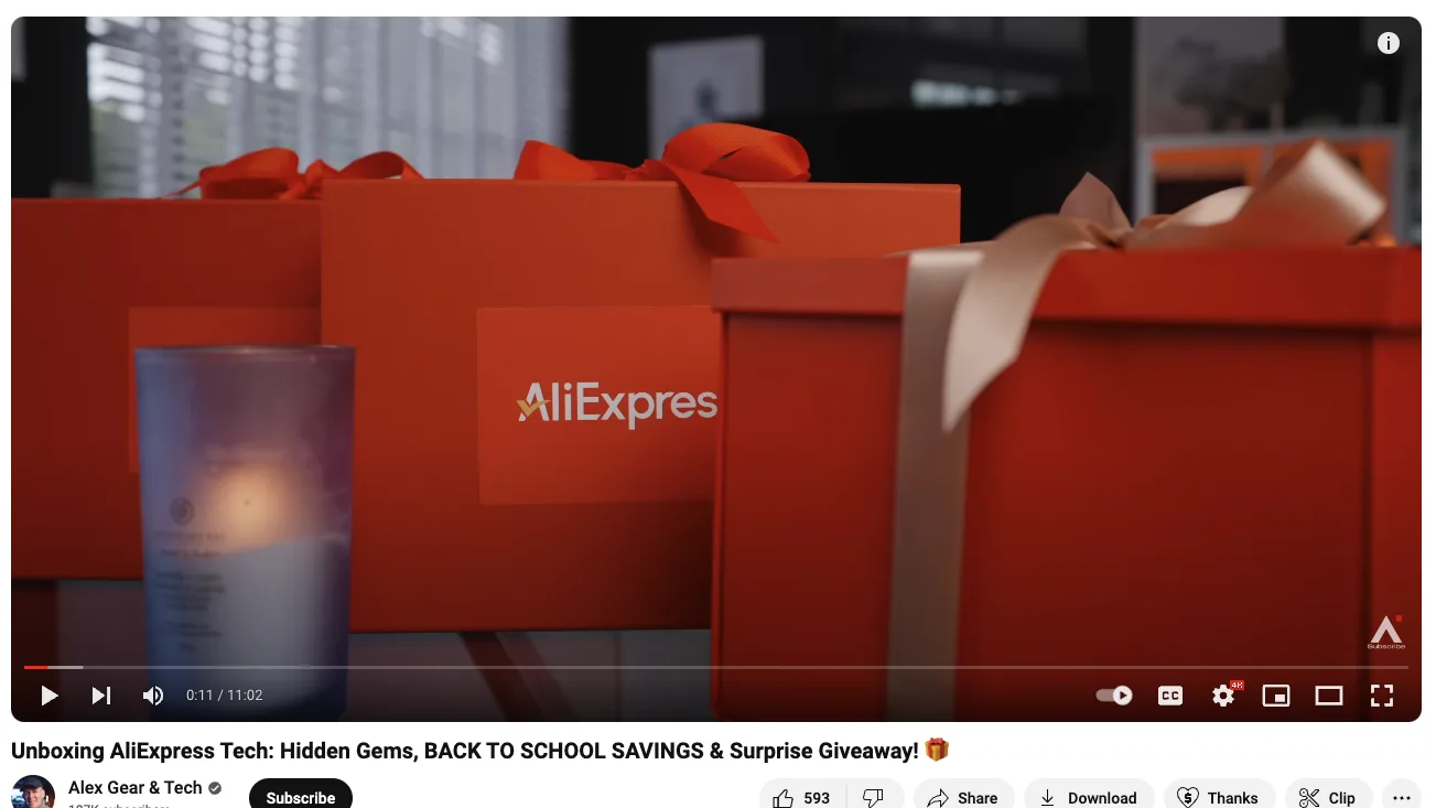 Video player still of orange AliExpress gift boxes with ribbons beside a lit candle, one box printed "AliExpres"