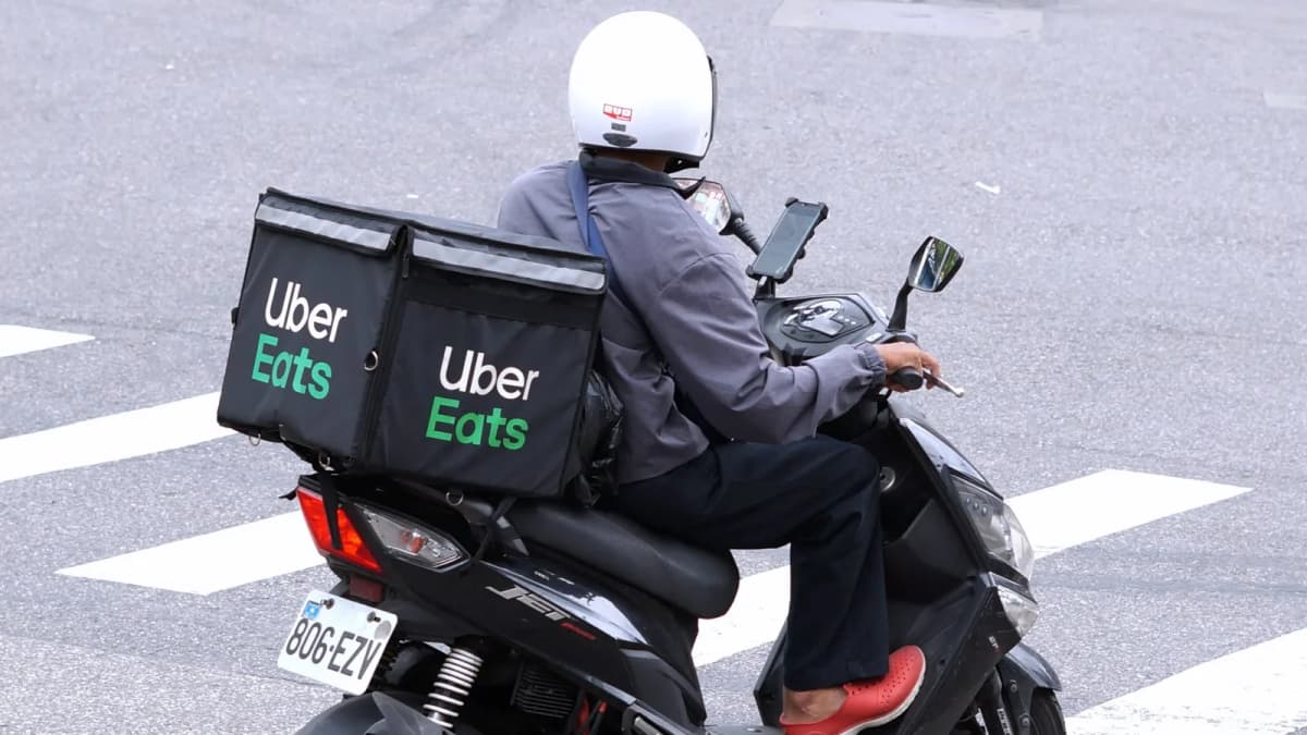 Delivery rider in a white helmet on a black scooter with an "Uber Eats" delivery box, crossing a street at a crosswalk
