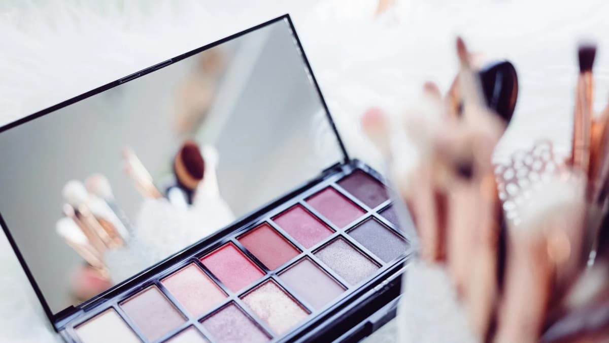 Open eyeshadow palette in pink and mauve shades with a mirrored lid, next to a blurred cup of makeup brushes