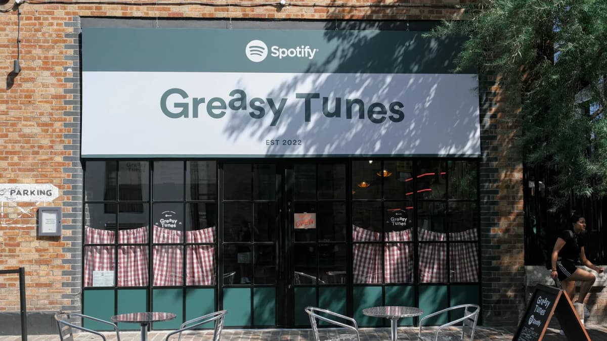 Brick cafe with a Spotify "Greasy Tunes" sign, gingham curtains and metal patio tables as a woman walks past