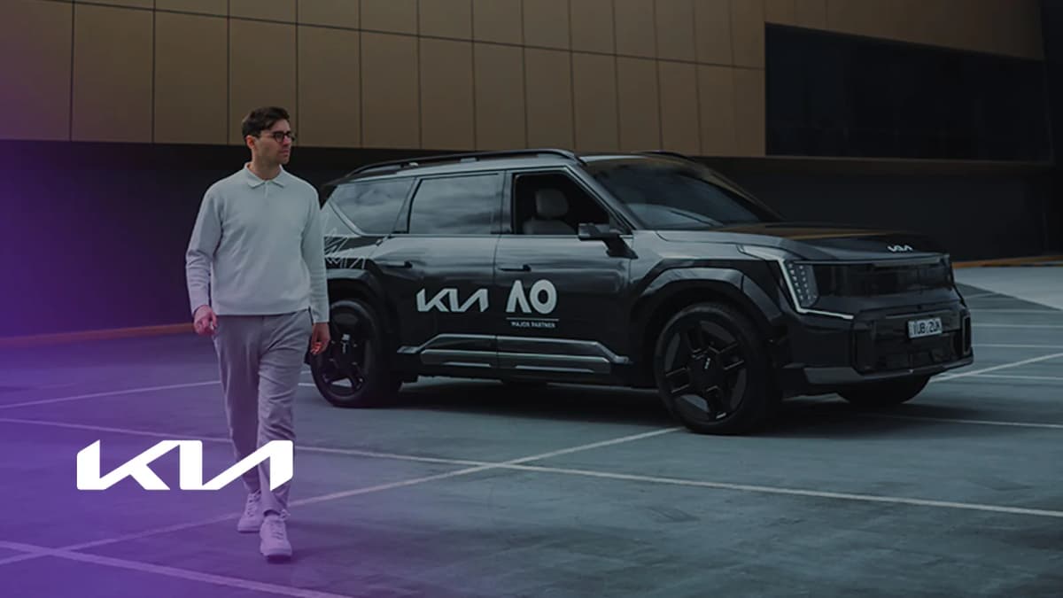 Man in glasses and a light sweater walks past a black Kia SUV marked "MAJOR PARTNER" in an empty car park