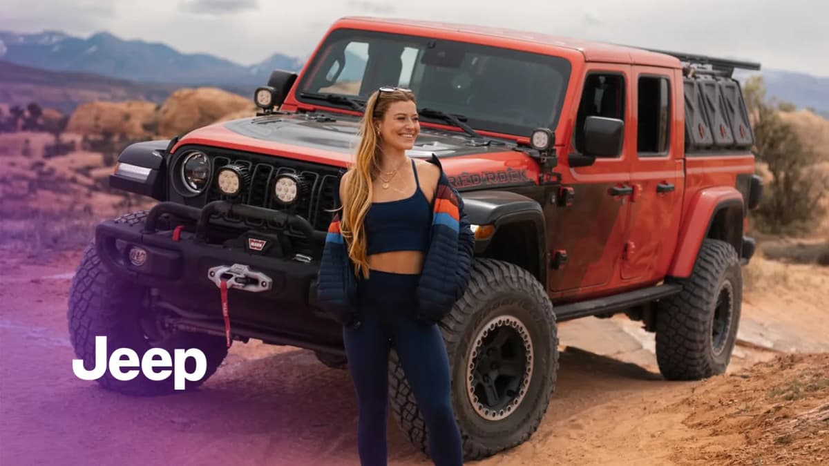Smiling woman with long hair stands before a red Jeep pickup marked "RED ROCK" on red desert rock with mountains behind