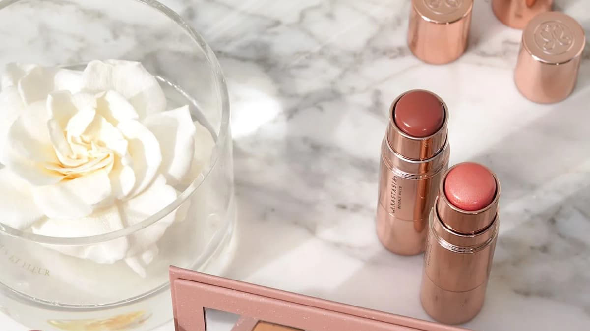 Rose-gold blush sticks printed "ANASTASIA BEVERLY HILLS" on marble beside a white flower in a glass and a pink palette