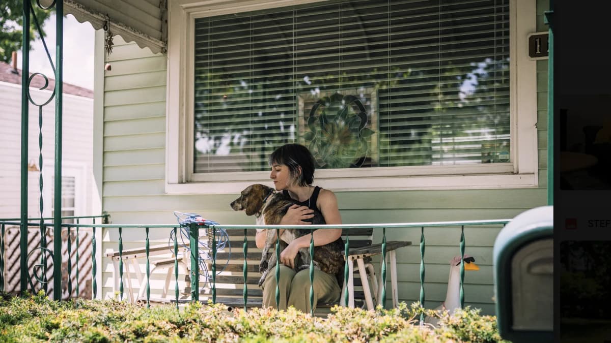 Woman with short dark hair sits on a bench on a green house's front porch, holding a beagle mix dog in her lap