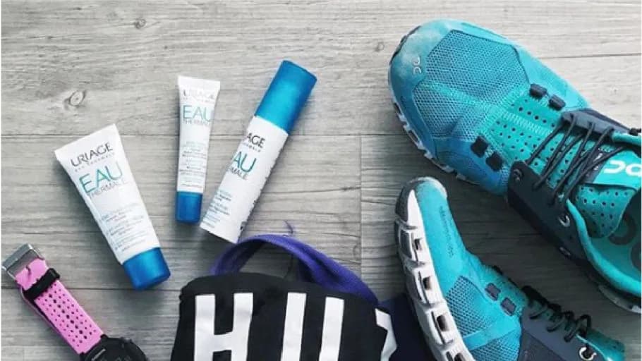 Three Uriage "EAU THERMALE" tubes on a wooden floor beside teal running shoes, a pink sports watch and a black bag