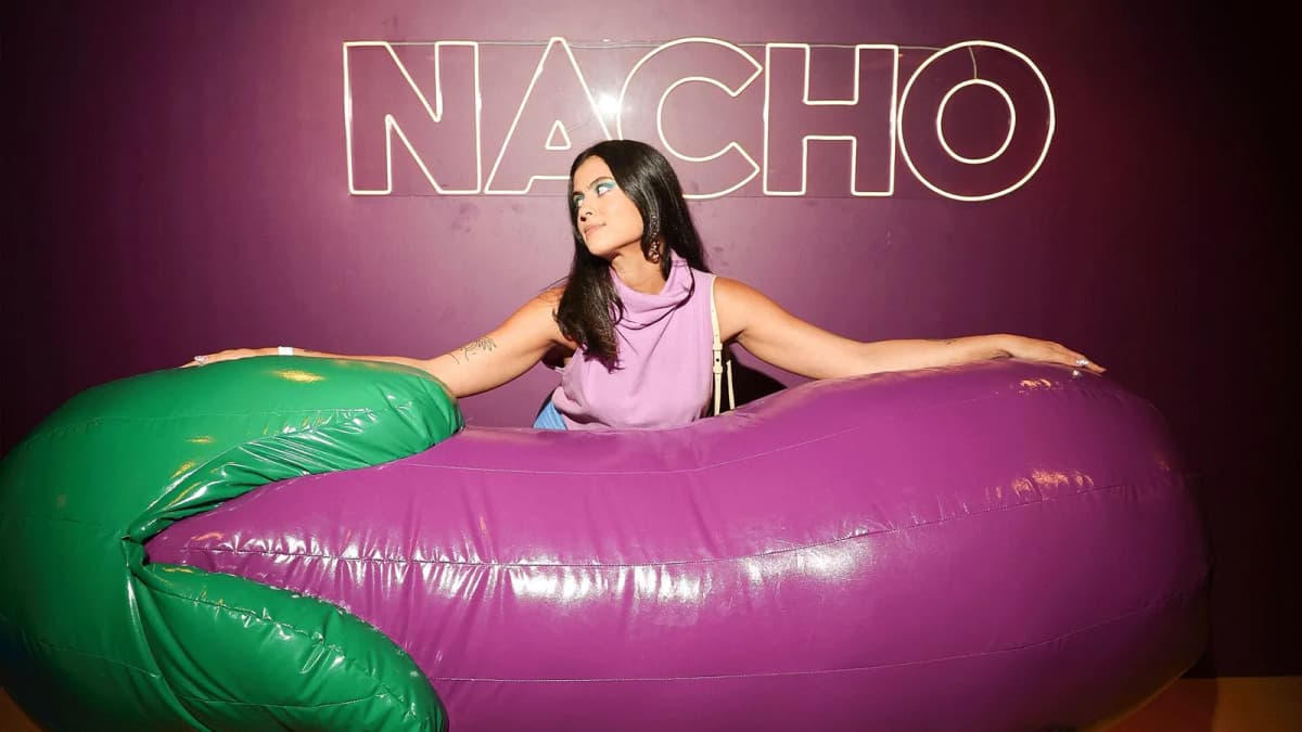 Woman in a lilac top leans on a giant inflatable eggplant beneath a neon "NACHO" sign on a purple wall