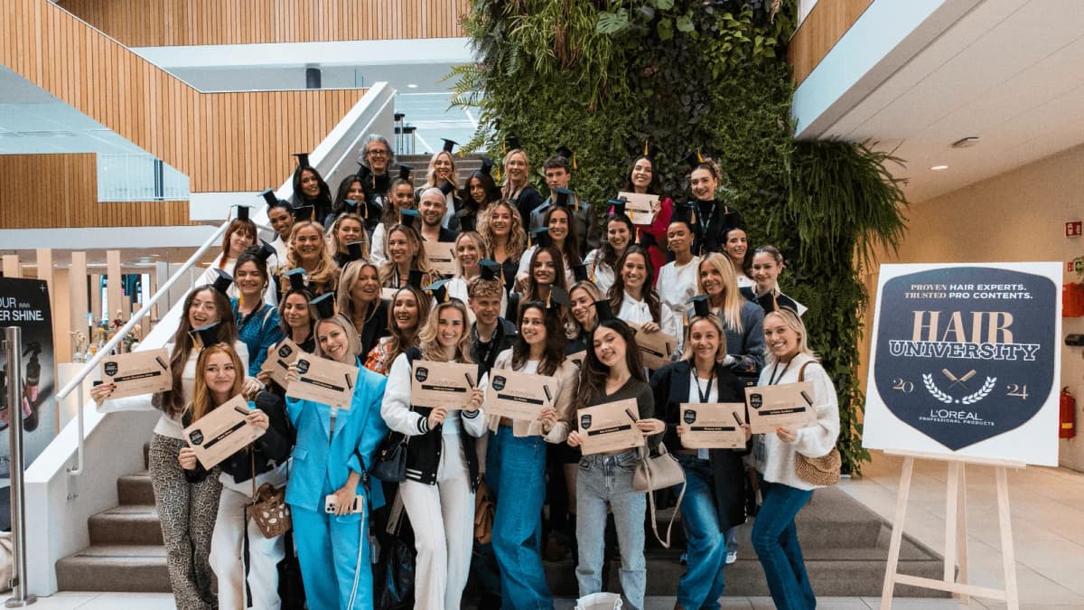 Group in graduation caps holding certificates on a staircase beside a L'Oréal Professional "HAIR UNIVERSITY" sign