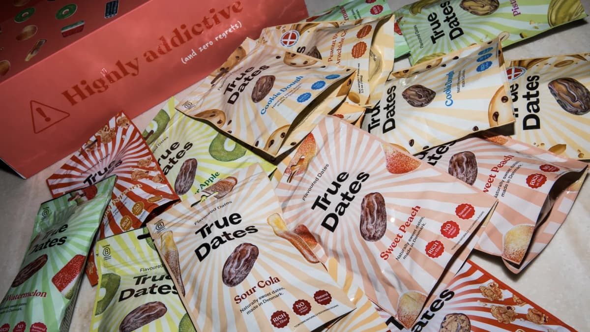 True Dates bags in flavours like "Sour Cola" and "Cookie Dough" spread on marble by a coral box reading "Highly addictive"