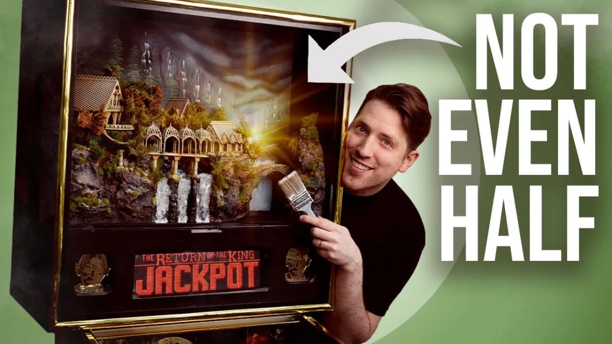 Smiling man holding a paintbrush beside a pinball backbox with a waterfall diorama, "JACKPOT" display, "NOT EVEN HALF"