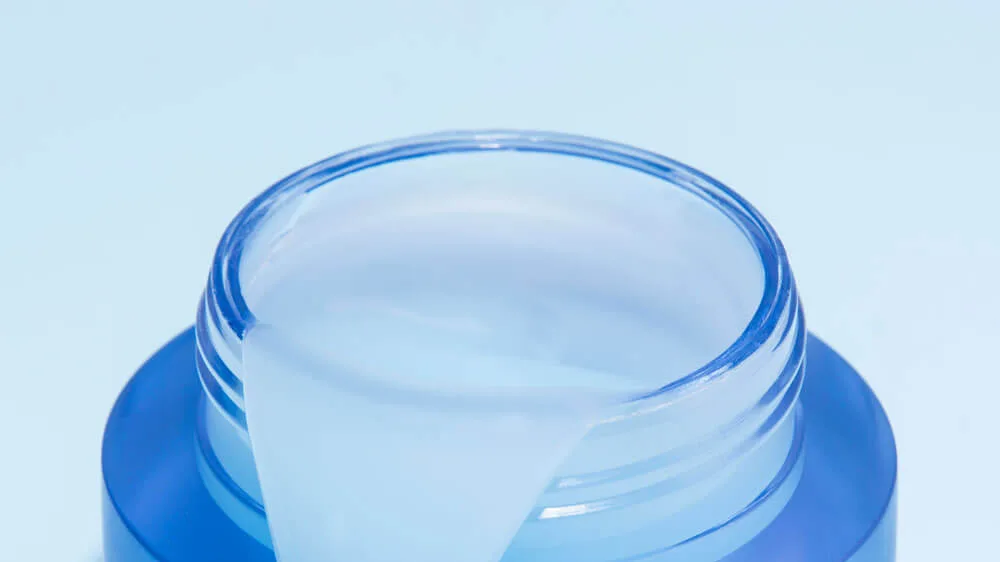 Close-up of an open blue glass jar filled with light blue gel cream against a pale blue background