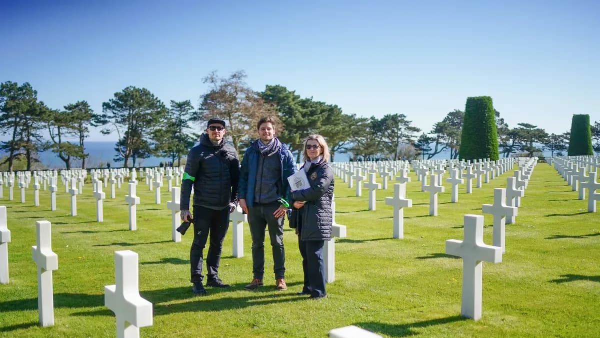 Two men and a woman in dark jackets stand among rows of white marble crosses in a seaside war cemetery on a sunny day