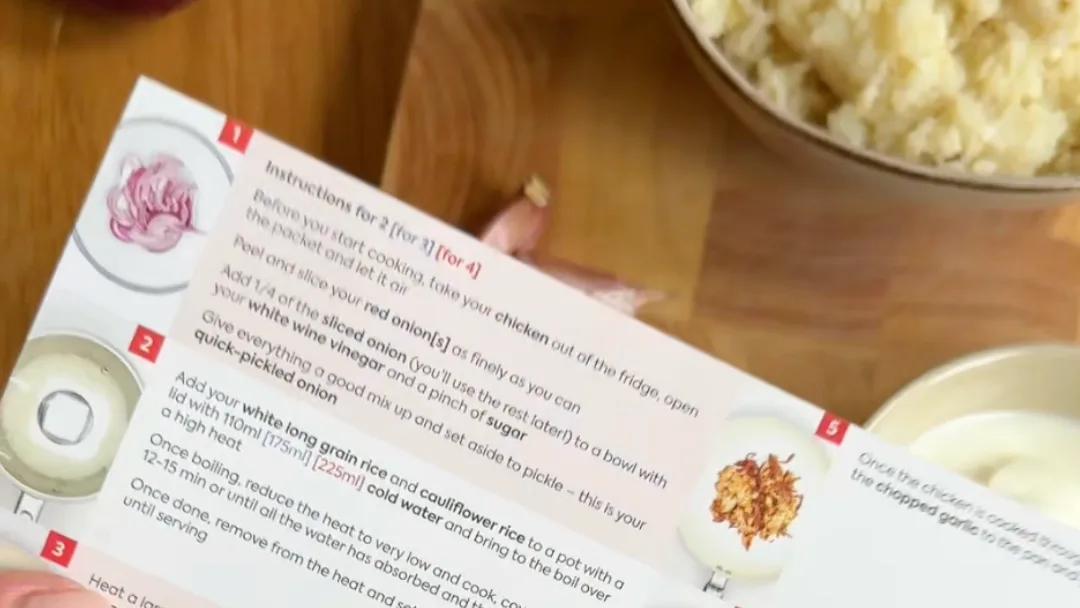 Recipe card reading "Instructions for 2" held over a wooden counter beside a bowl of cooked rice and a garlic clove