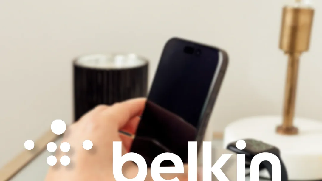Hand holding a smartphone near a candle, brass lamp and smartwatch on a bedside table, with the Belkin logo overlaid