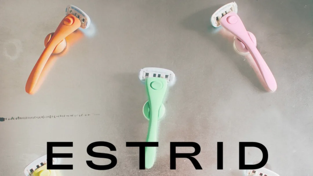Orange, green and pink Estrid razors on suction holders against a grey wall, with "ESTRID" printed across the bottom