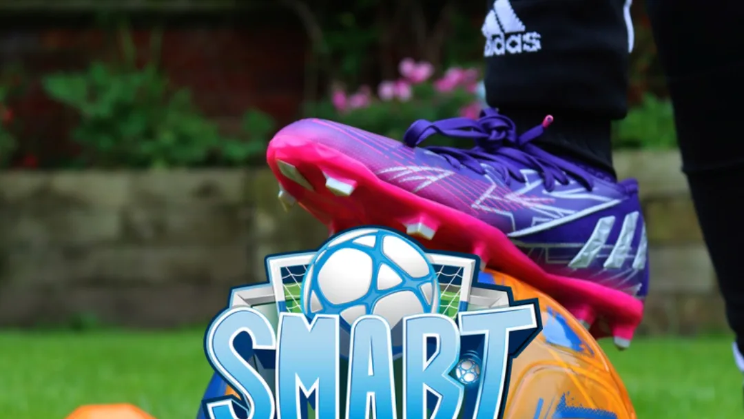Foot in a pink and purple football boot rests on an orange ball on grass, with the Smart Ball logo reading "SMART"