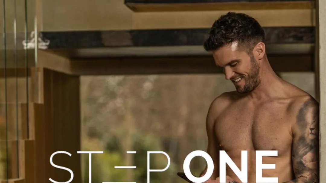 Shirtless, smiling man with a tattooed arm looks down at a phone beside a staircase, with a "STEP ONE" logo across it