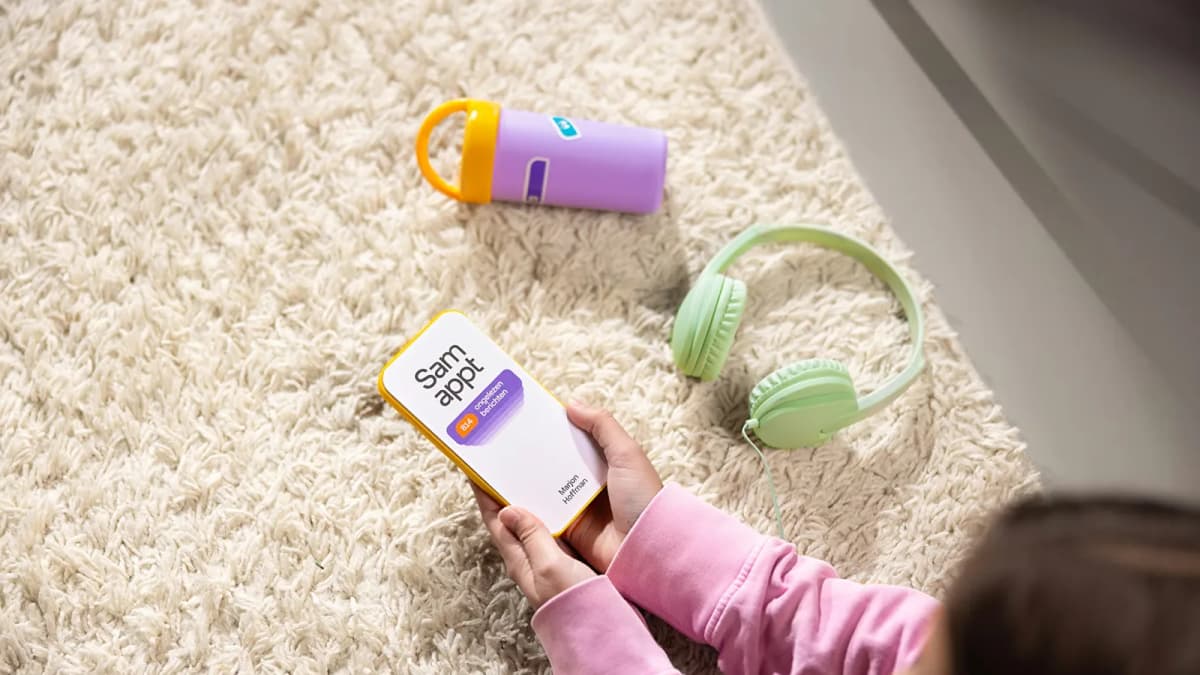 Child in pink sweater holds phone-shaped book "Sam appt" on shaggy rug beside green headphones and purple bottle