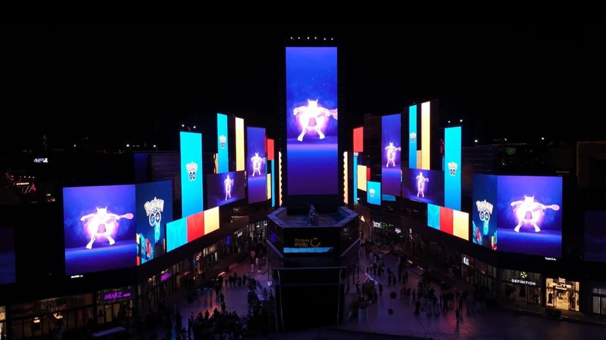 Night aerial view of a plaza lined with giant screens showing Mewtwo and the Pokémon GO logo as crowds walk below
