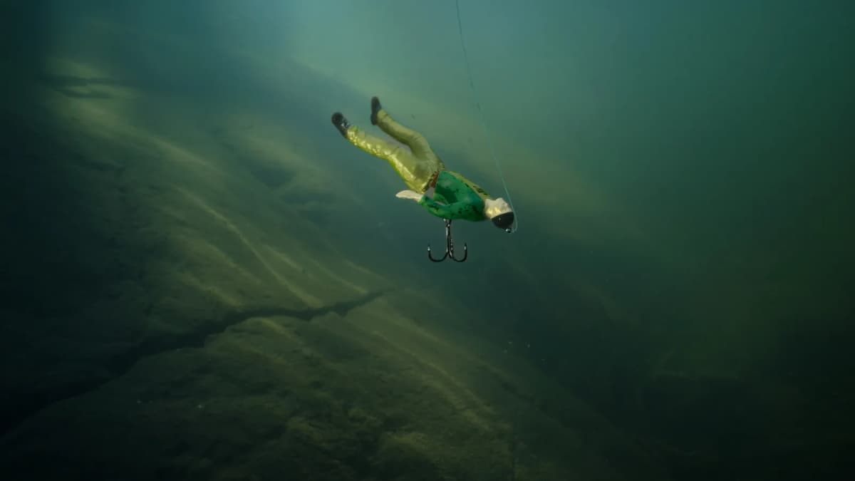 Fishing lure shaped like a man in a green shirt and khaki trousers, with a treble hook, sinking on a line in murky water